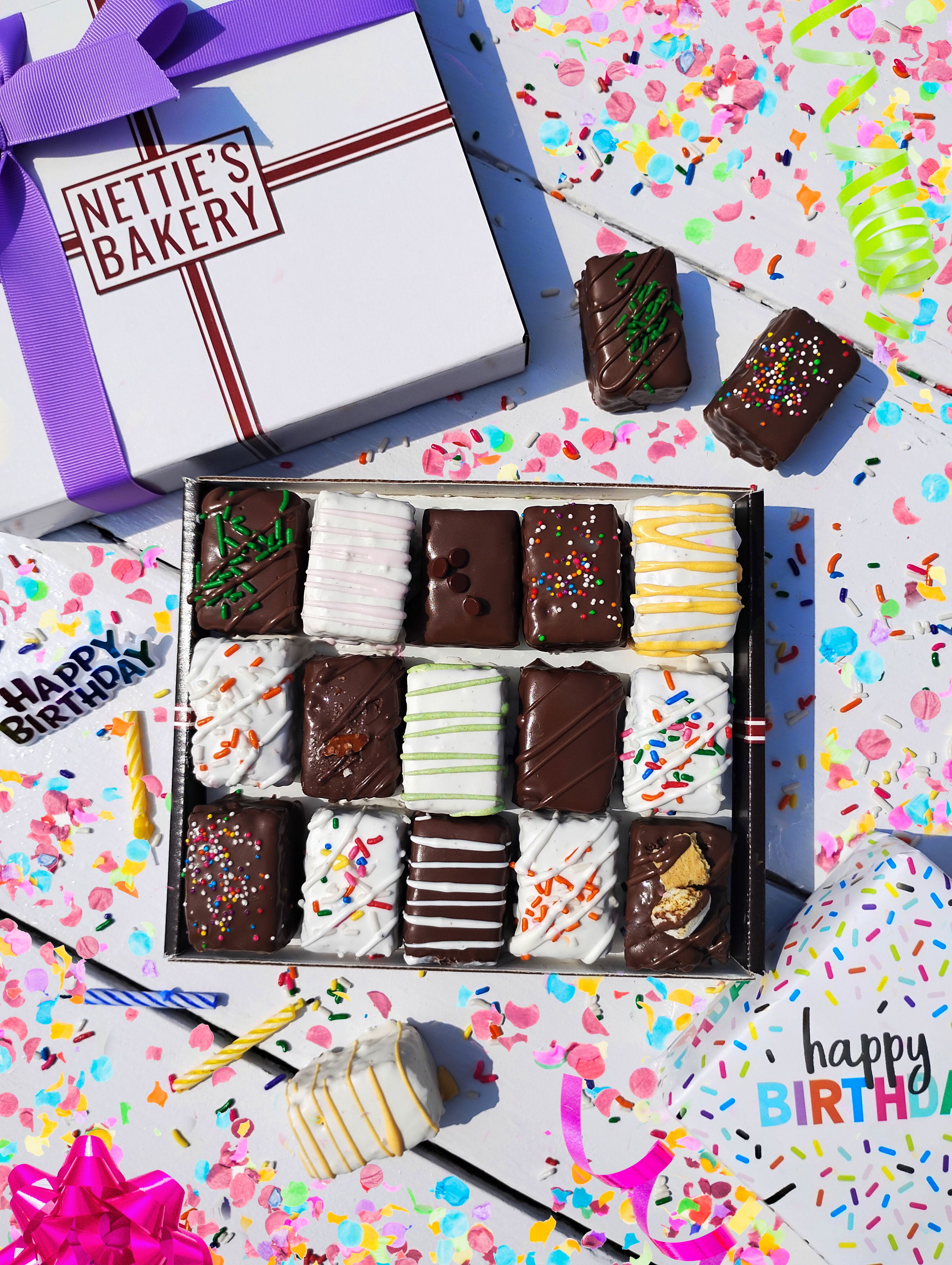 A box of brownie and cake bites on a white surface surrounded by birthday decorations.