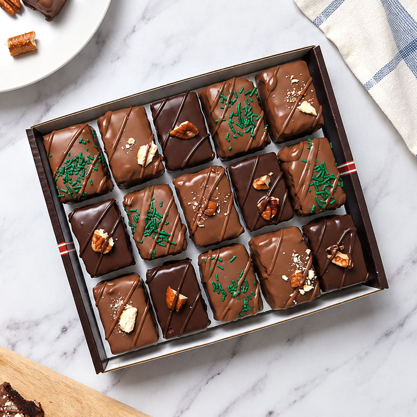 Open gift box of 15 chocolate-covered brownie bites in assorted boozy-inspired flavors, styled with a plate and serving board on white marble.