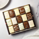 Nettie’s Bakery Cake Combo gift box with 15 chocolate-covered cake bites in white and milk chocolate coatings, finished with colorful sprinkles and pastel drizzles. A plate beside the box shows several cut cake bites and their layered centers.
