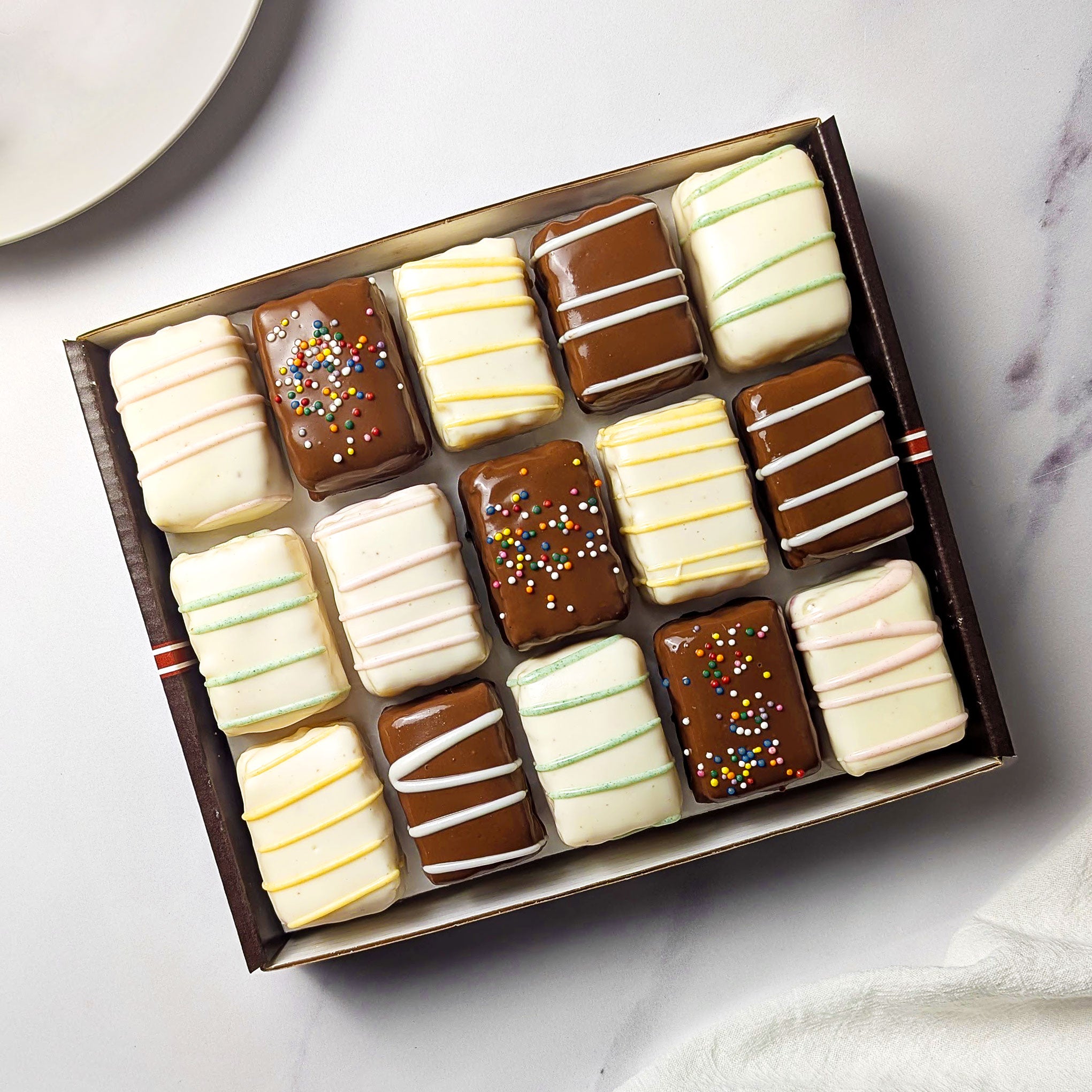 Nettie’s Bakery Cake Combo gift box with 15 chocolate-covered cake bites in white and milk chocolate coatings, finished with colorful sprinkles and pastel drizzles. A plate beside the box shows several cut cake bites and their layered centers.