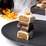 Nettie’s Bakery Halloween treats: white chocolate-covered Candy Corn Blondie Bites and a chocolate-covered Reese’s Pieces Brownie Bite, cut open and stacked on a charcoal plate, with a gift box and Halloween decorations in the background.