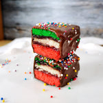 Two rainbow cookies sitting on a white surface with a rustic background. The rainbow cookies are stacked and cut in half to show the inside. Gift-ready and shipped nationwide.