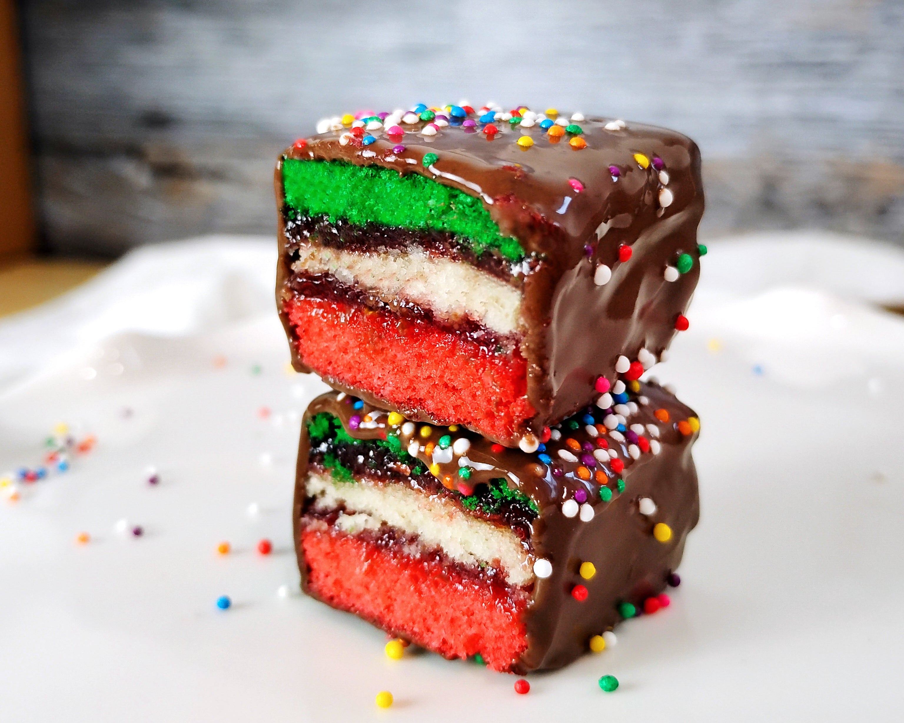 Two rainbow cookies sitting on a white surface with a rustic background. The rainbow cookies are stacked and cut in half to show the inside.