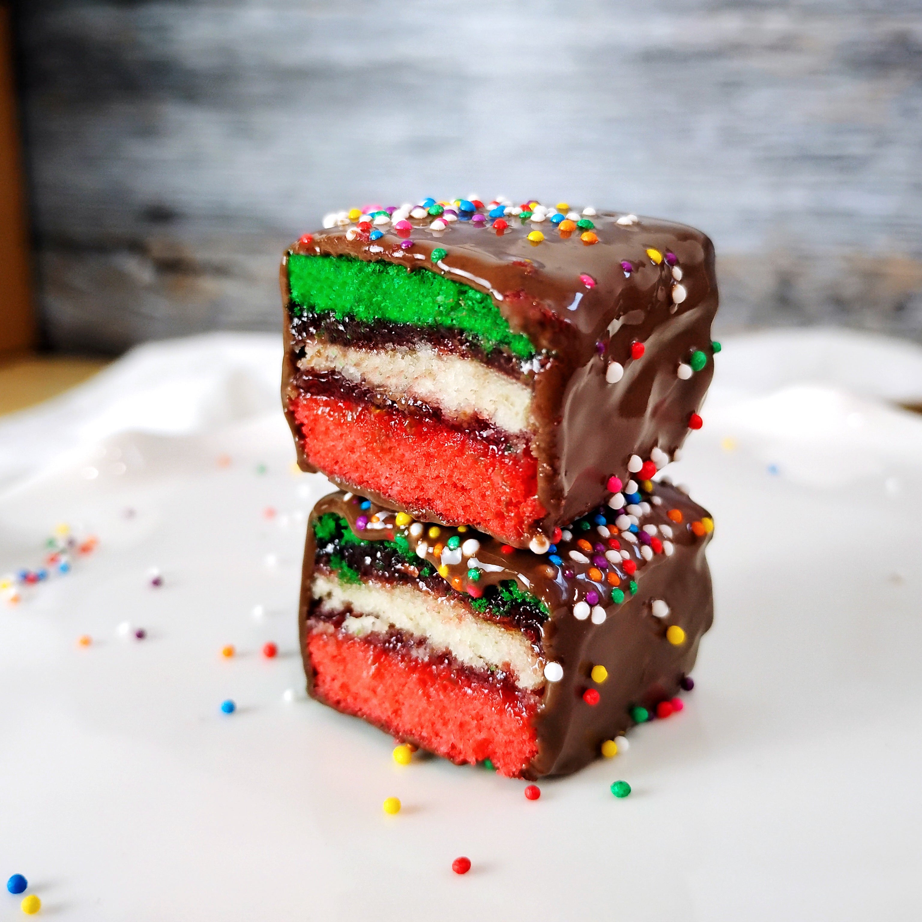 Two rainbow cookies sitting on a white surface with a rustic background. The rainbow cookies are stacked and cut in half to show the inside.