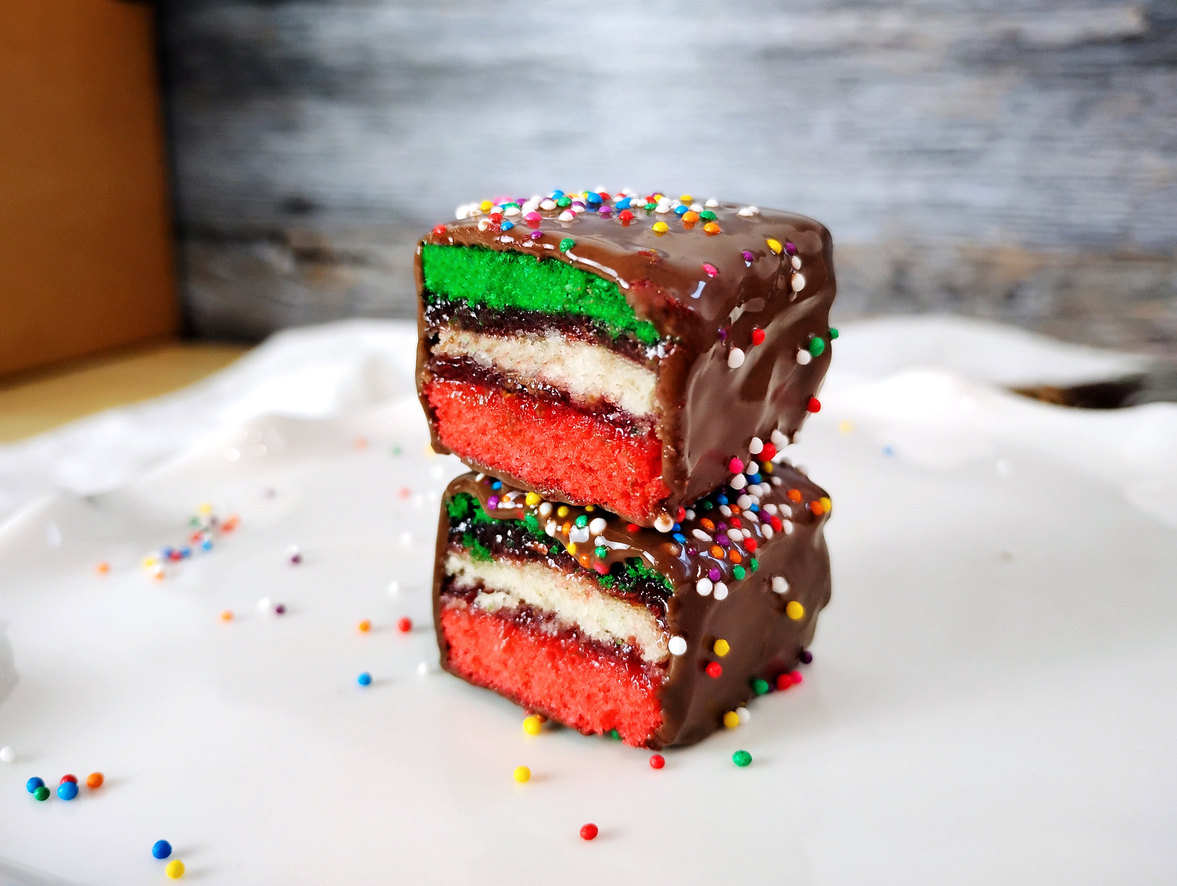 Two rainbow cookies sitting on a white surface with a rustic background. The rainbow cookies are stacked and cut in half to show the inside.